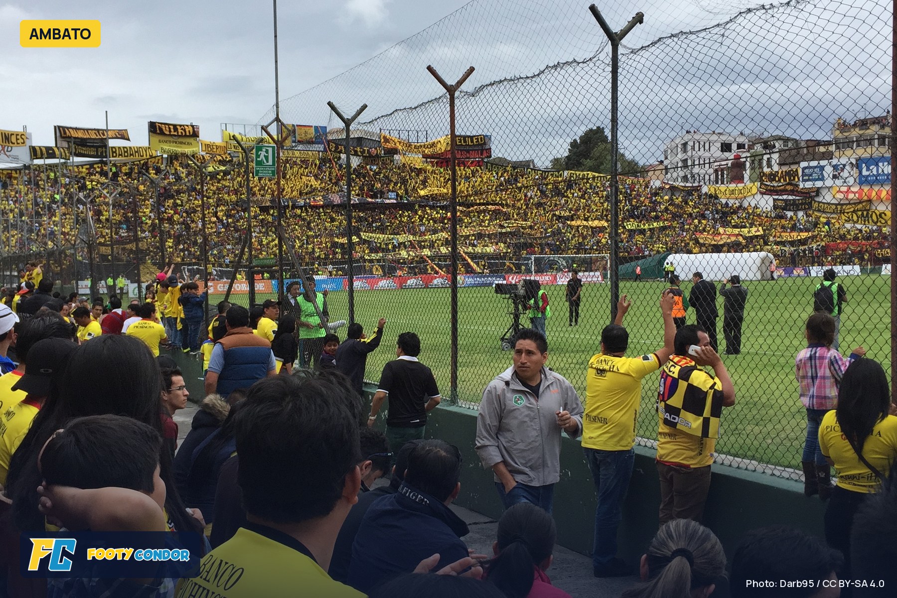 Packed stands at Estadio Bellavista in Ambato, seen from behind the fence, with fans in yellow